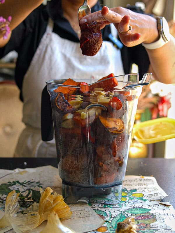 A person wearing an apron prepares to blend a variety of colorful ingredients including fruits, vegetables, and spices in a blender. They hold a spoon above the blender, suggesting an addition of gochujang. Nearby are corn husks and paper.